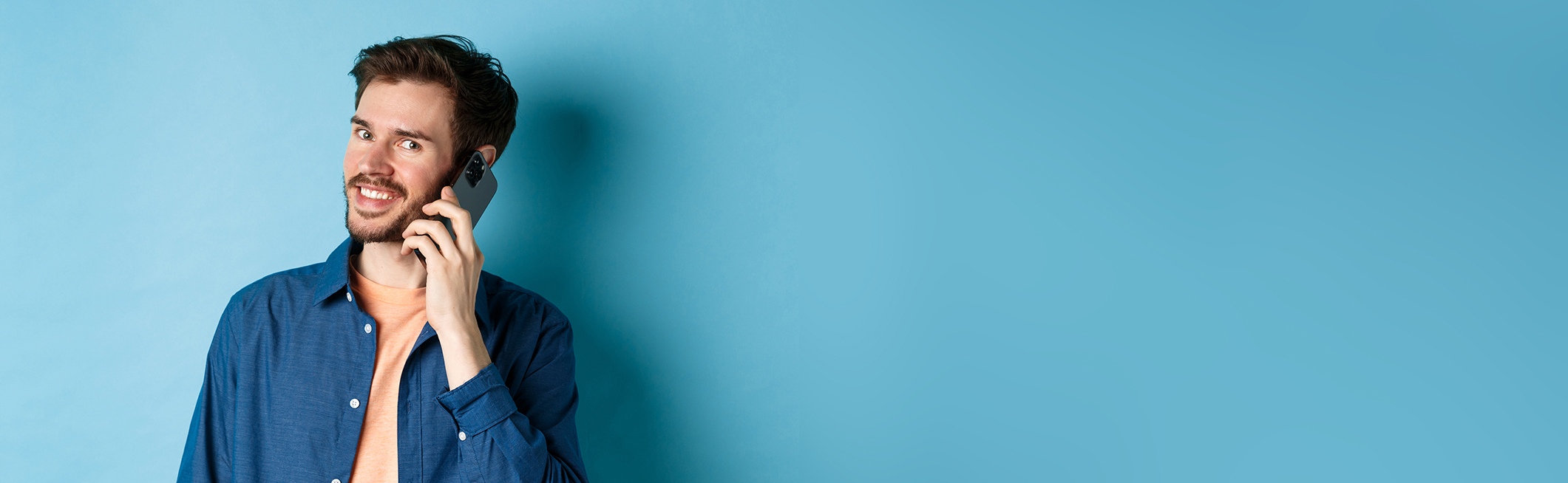 Modern smiling guy talking on mobile phone, looking happy at camera, standing on blue background.